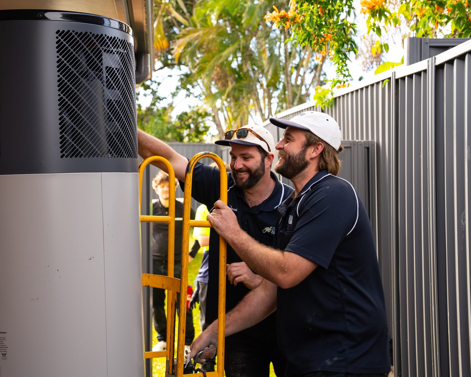 Two Goanna plumbers installing a Rinnai heat pump hot water system beside a brick home